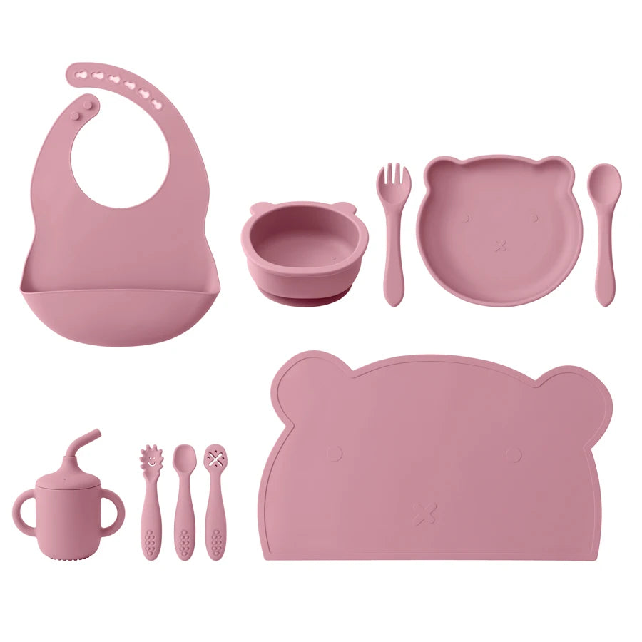 Pink children's dining set including bib, bowl, plate, cup, and utensils on a white background