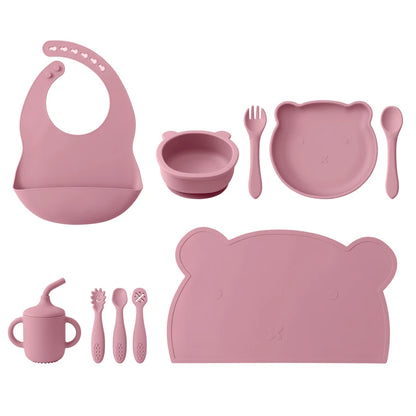 Pink children's dining set including bib, bowl, plate, cup, and utensils on a white background