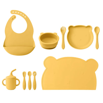 Yellow silicone baby feeding set including bib, bowl, plate, spoon, fork, cup, and placemat on a white background.